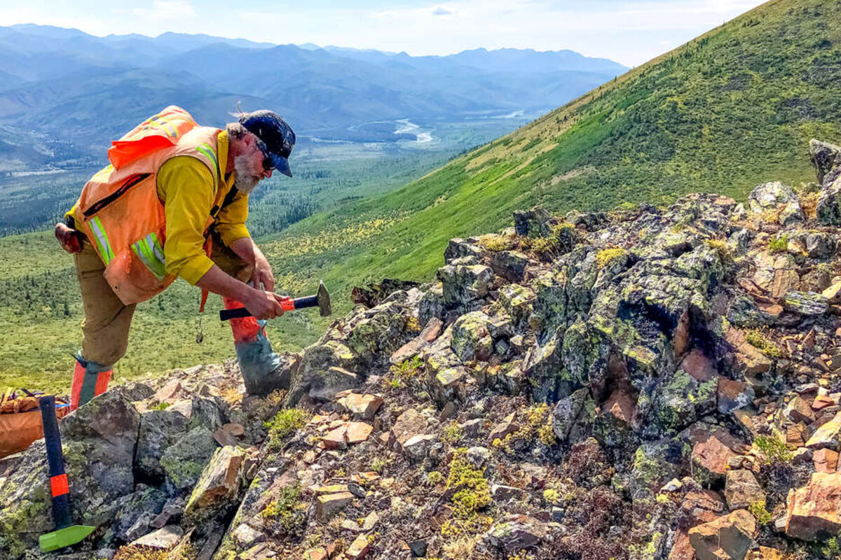 Geologist uses hammer to break a critical minerals sample off a rock in Alaska.
