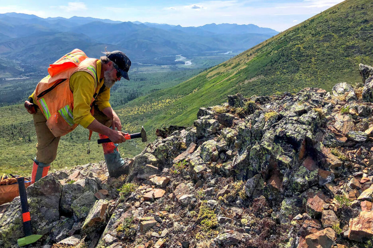 Geologist using hammer to break off a rock sample on a hillside in Alaska.