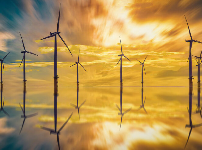 A sunset paints the sky orange behind large offshore wind turbines.