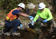 Two men in safety gear collect water samples from a stream.