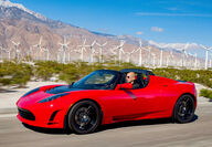 A red Tesla Roadster speeds past a large wind turbine farm.