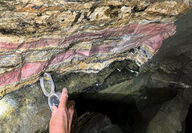 A hand holds an instrument against bands of pink and cream mineralization.