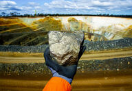 Extended hand holding a large chunk of nickel ore with a mine in the background.