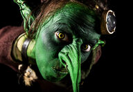 Closeup of the face of green goblin with a miner’s light strapped to its head.